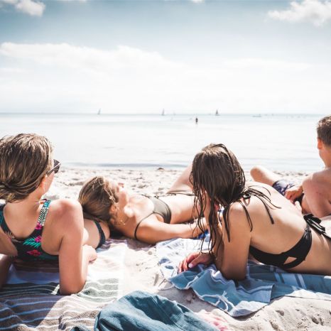 Rückansicht einer Freundesgruppe aus drei Frauen und einem Mann liegt teilweise aneinandergelehnt im Sand und schaut auf's Meer