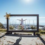 Frau in weißen Kleid steht im Fotorahmen oberhalb des Yachthafens, im Hintergrund die Ostsee, der Yachthafen und strahlender Sonnenschein