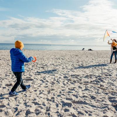 Frau und Kind lassen gemeinsam einen Drachen steigen. Das Kind ist von hinten zu sehen und hält die Leinen, die Frau ist im Hintergrund von vorn zu sehen und gibt dem Drachen Starthilfe
