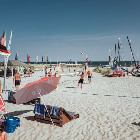 Blick auf das Treiben am Strand der Surfschule: Im Vordergrund Sitzgelegenheiten, dahinter ein bespieltes Beachvolleyballfeld und im Hintergrund Flaggen, Masten, Segel und die Ostsee