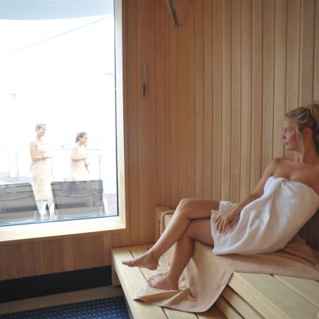 Frau sitzt in Handtuch gewickelt in einer Sauna neben großem Fenster mit Blick auf zwei Frauen draußen