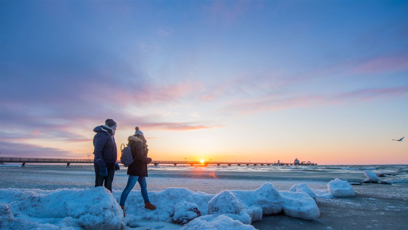 Zwei Personen in Winterkleidung gehen am eisbedeckten Strand bei Sonnenuntergang entlang