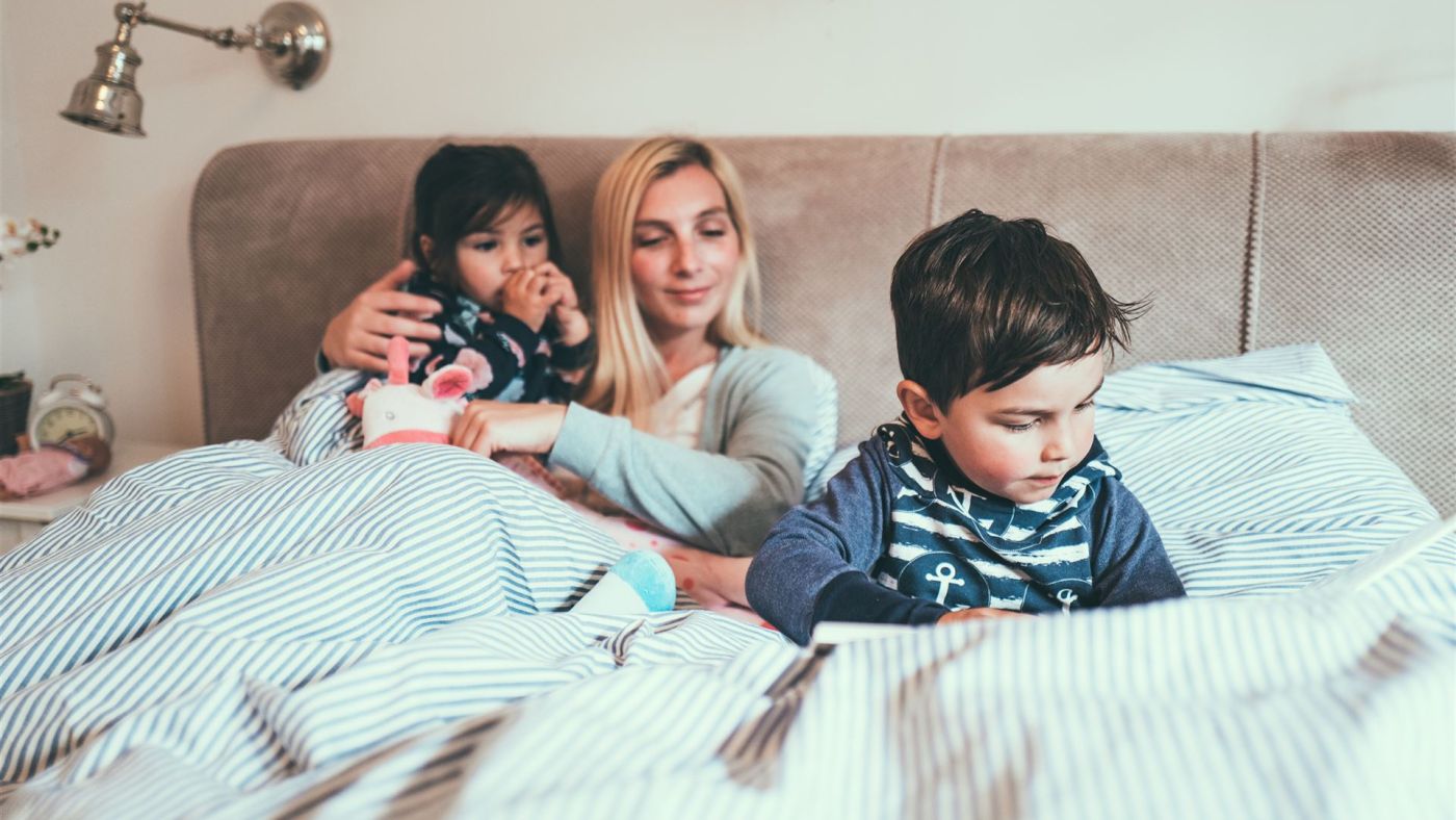 Frau sitzt mit zwei Kindern im Bett, eines hält ein Kuscheltier, anderer schaut nach unten