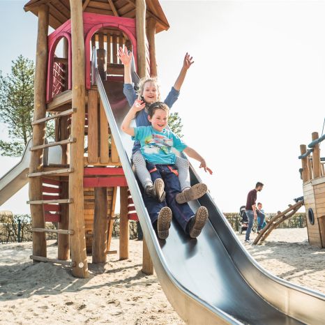 Zwei Kinder genießen eine Rutschpartie auf einem Spielplatz mit Holzschaukel und Sand unter den Füßen.